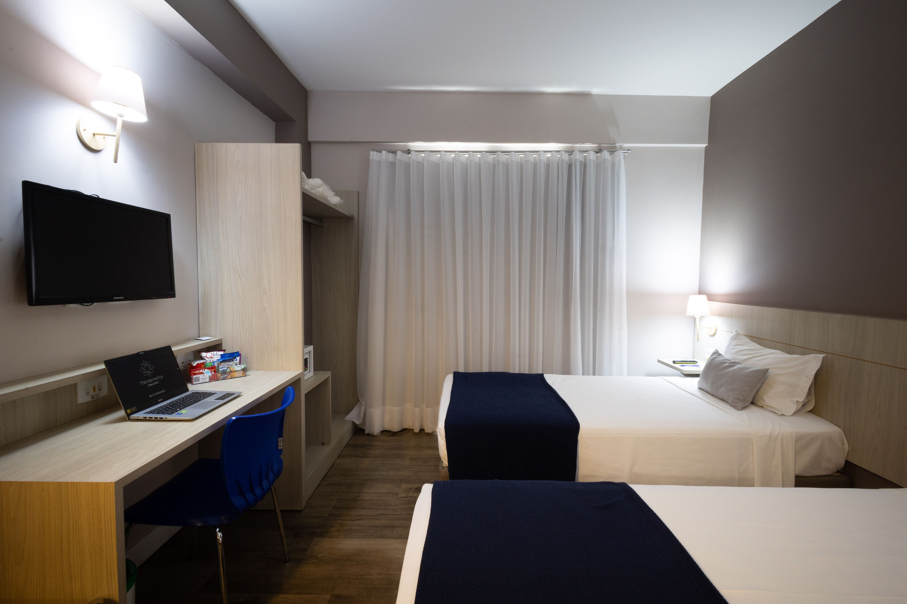 A hotel room with a desk, two beds, a TV, and a shower area.