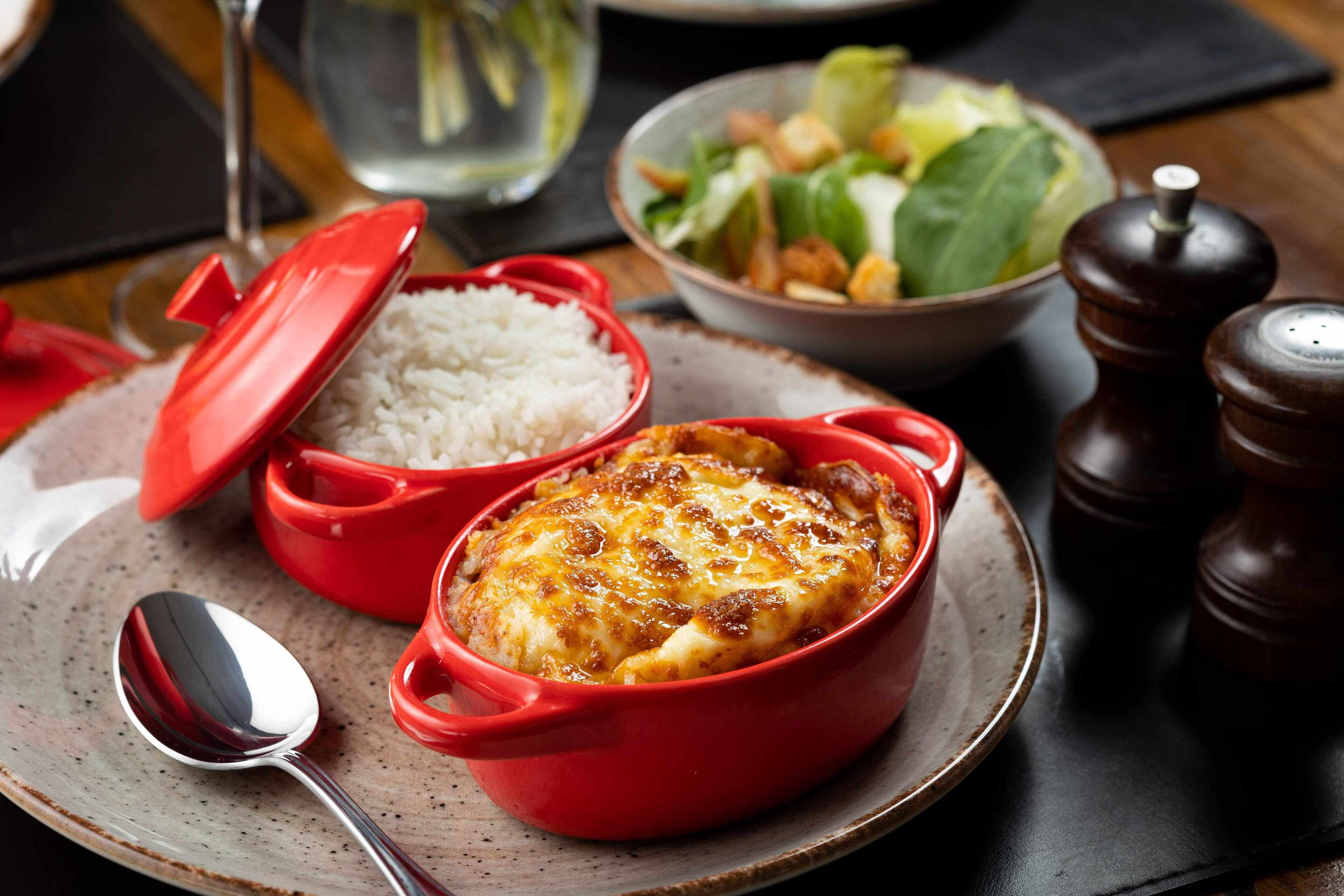A meal with rice and a baked dish in red pots, a salad, and a pepper grinder on a table.