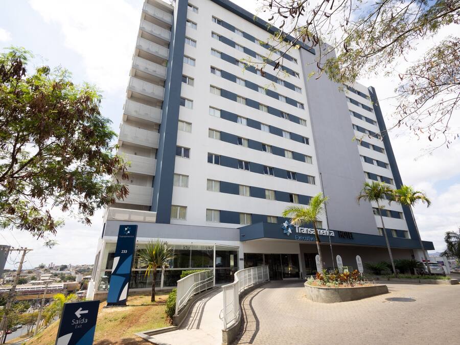 Um hotel moderno com vários andares, entrada coberta e área ajardinada com palmeiras.