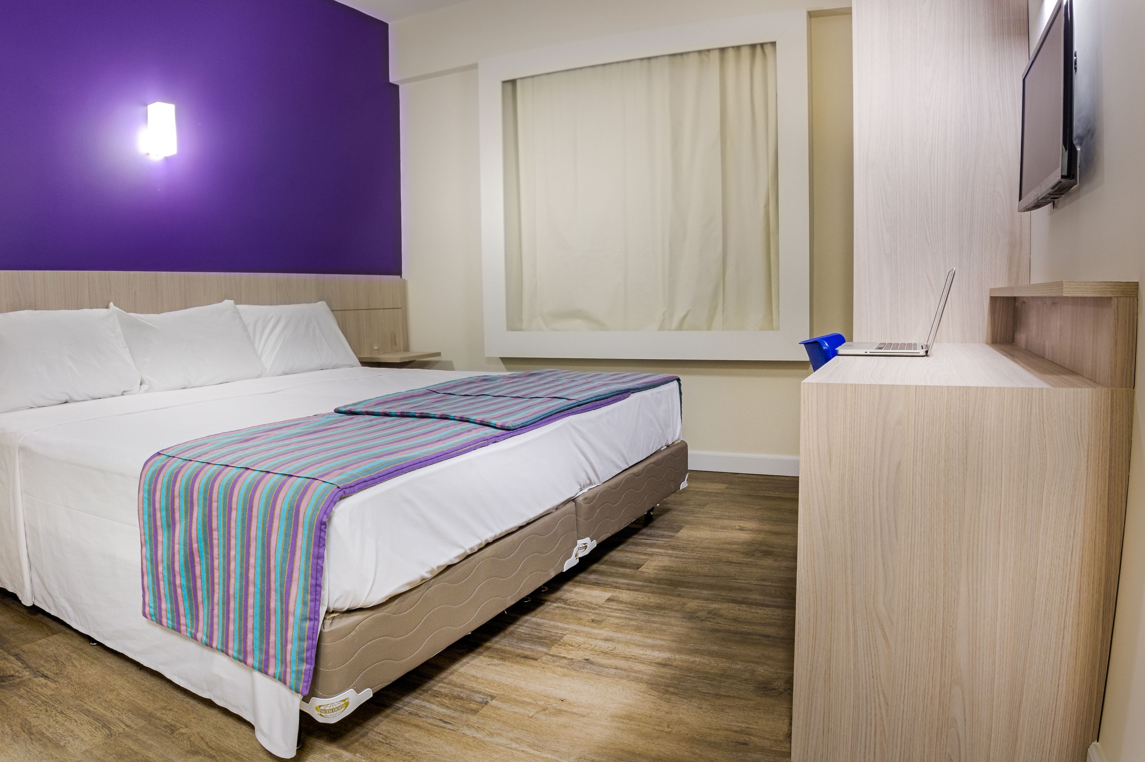 A hotel room with a bed, a desk, a laptop, a window with curtains, and a TV on the wall.