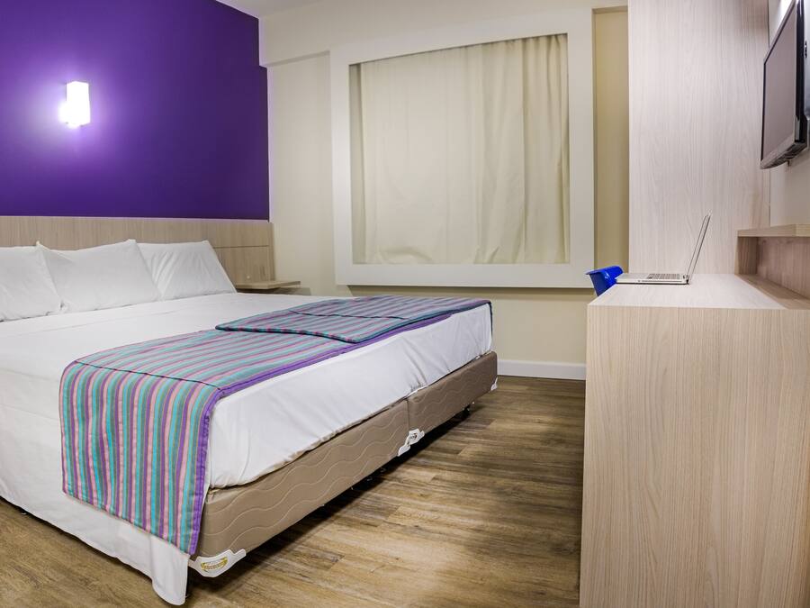 Habitación de hotel con cama, escritorio, computadora portátil, ventana con cortinas y televisor en la pared.