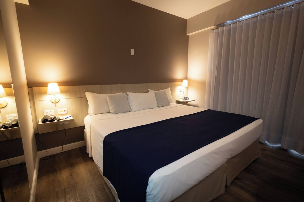 A hotel room with a large bed, bedside lamps, a telephone, and a window with curtains.