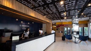 Un área de recepción moderna con techo de madera y un mostrador negro. Hay dos recepcionistas detrás del mostrador, y una zona de estar con sillas azules y una mesa al fondo.