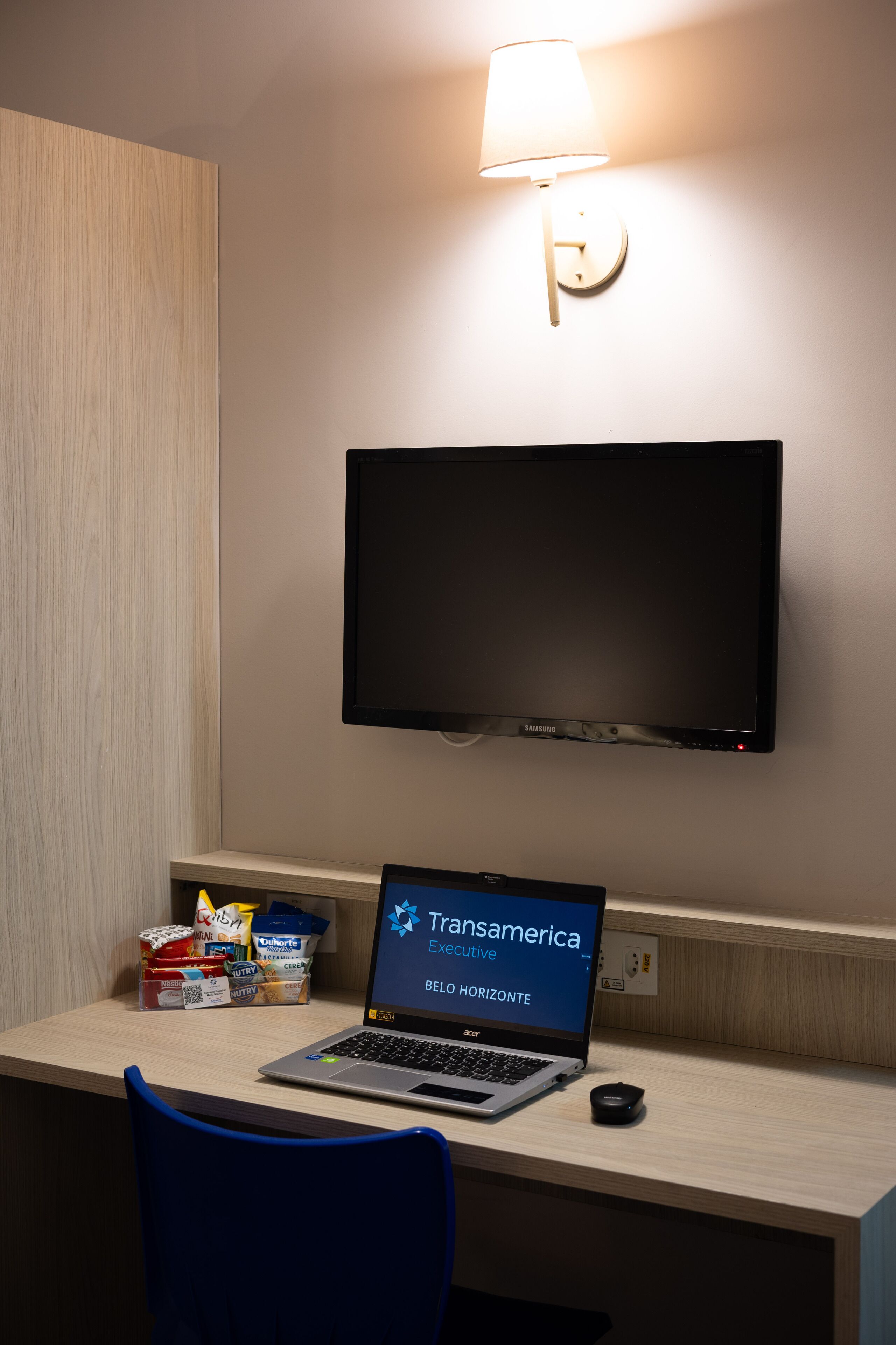 A hotel room with a desk, a laptop, a television, and a wall-mounted lamp.