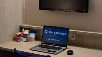 A hotel room with a desk, a laptop, a television, and a wall-mounted lamp.
