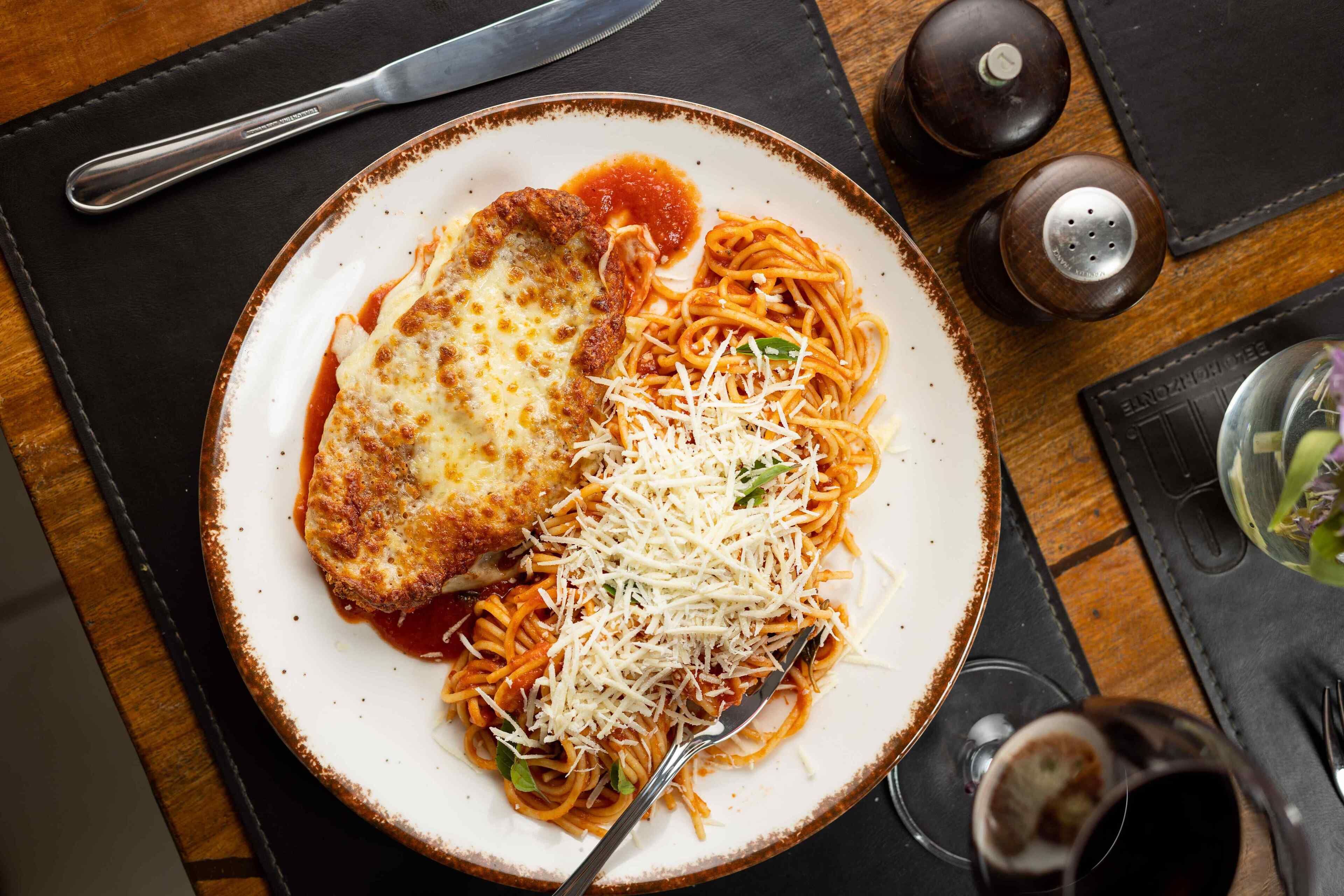 Een bord spaghetti met een stukje gepaneerde kip en geraspte kaas op een tafel, samen met een mes, zout- en peperstrooiers en een glas water.