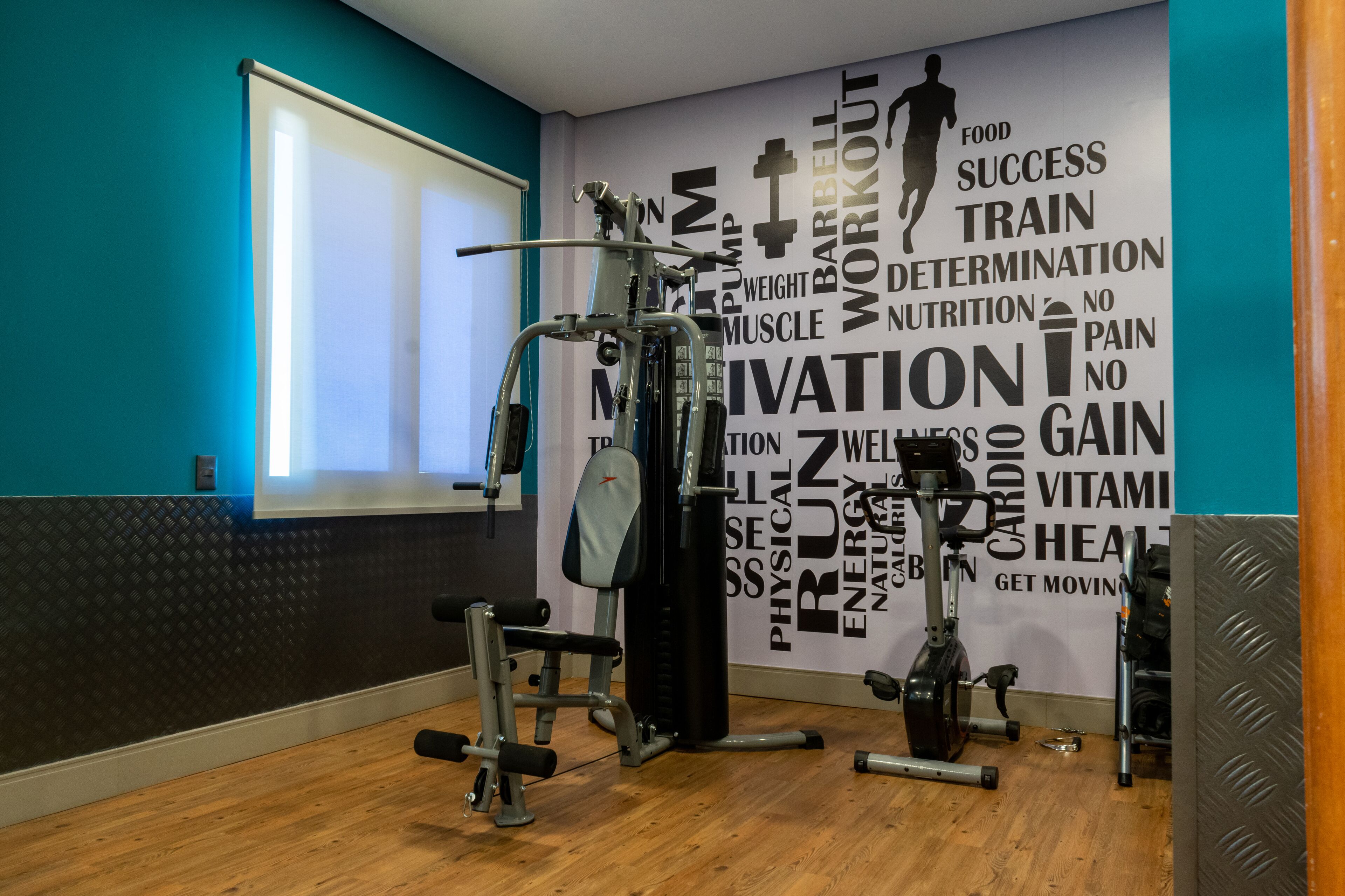 Un gimnasio con diverso equipo de ejercicio, una pared con palabras motivacionales y una ventana con persianas.