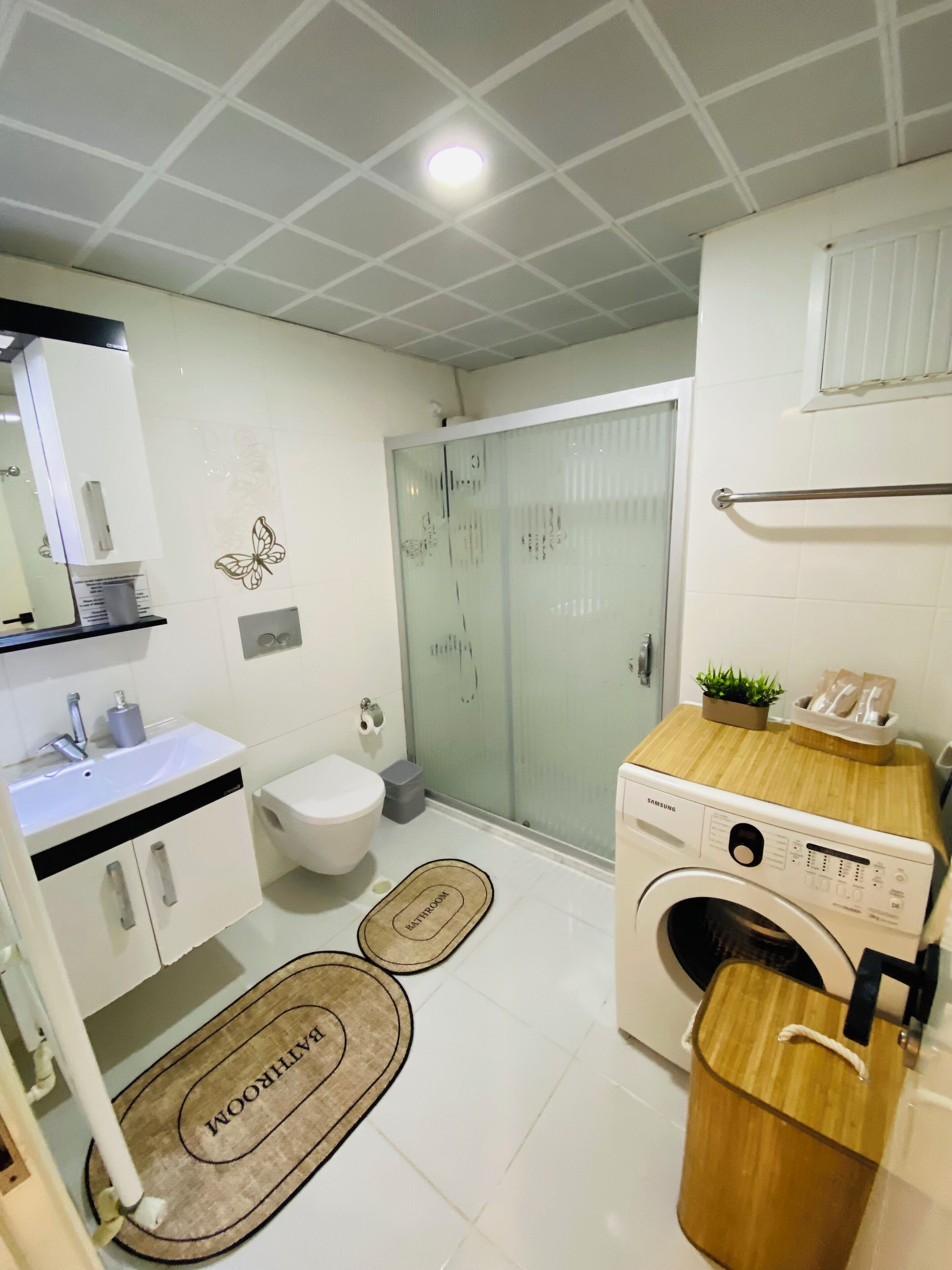 Ein kleines Badezimmer mit weißem Waschbecken, Toilette und Waschmaschine, einer Duschkabine aus Glas und einer Holzablage mit einer kleinen Pflanze.