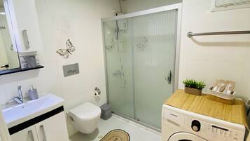Ein kleines Badezimmer mit weißem Waschbecken, Toilette und Waschmaschine, einer Duschkabine aus Glas und einer Holzablage mit einer kleinen Pflanze.