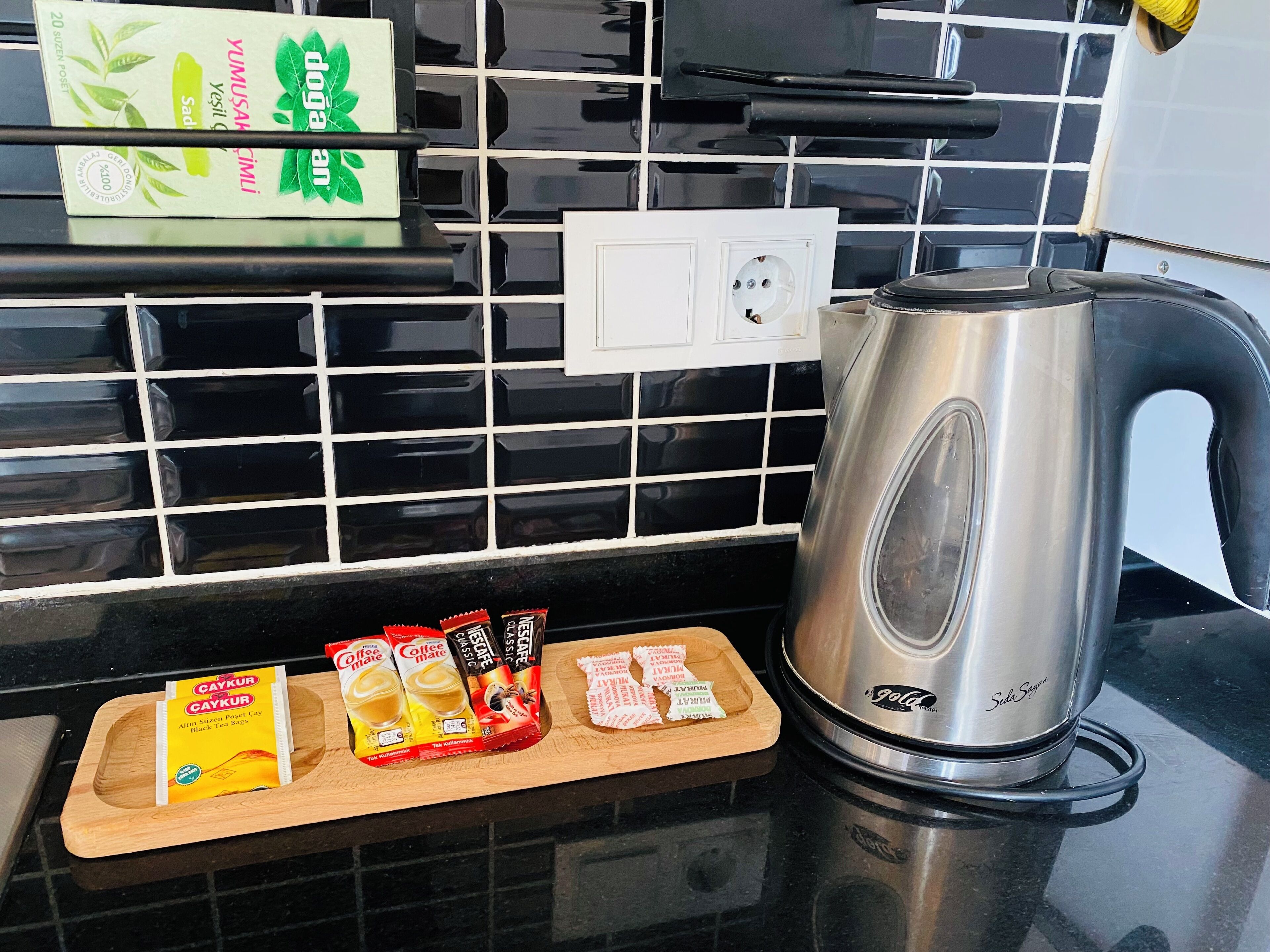 A kitchen counter with a stainless steel kettle, a wooden tray with tea bags, and a box of black tea.