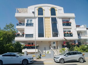 A multi-story building with balconies, a central entrance, and cars parked in front.
