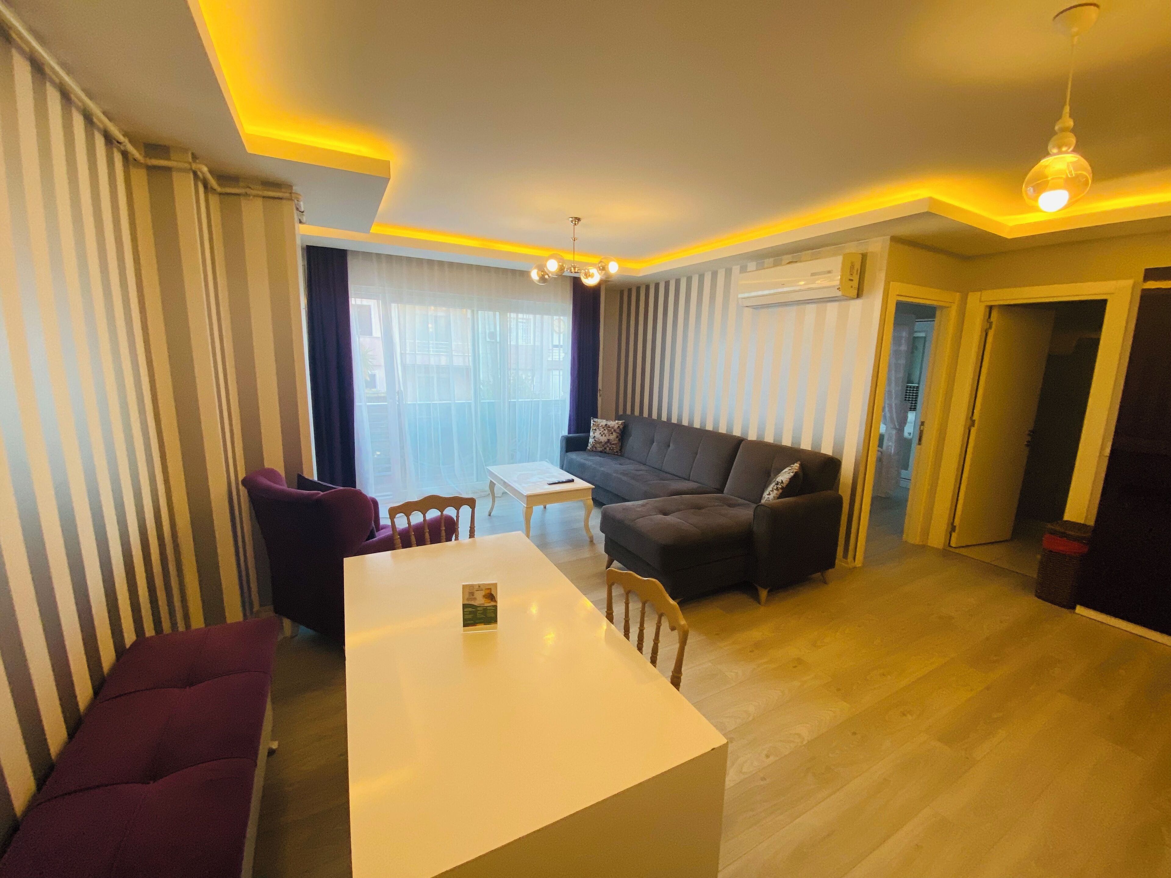 A modern living room with a sofa, a dining table, and a chair. There is a window with curtains, a ceiling light, and an air conditioning unit.