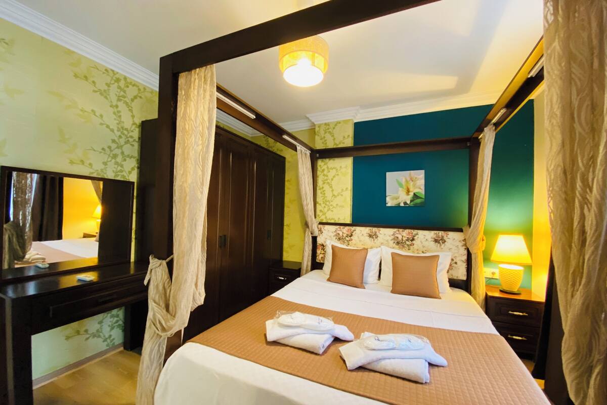 A four-poster bed with a headboard, a bedside table, a mirror, and a nightstand with a lamp.