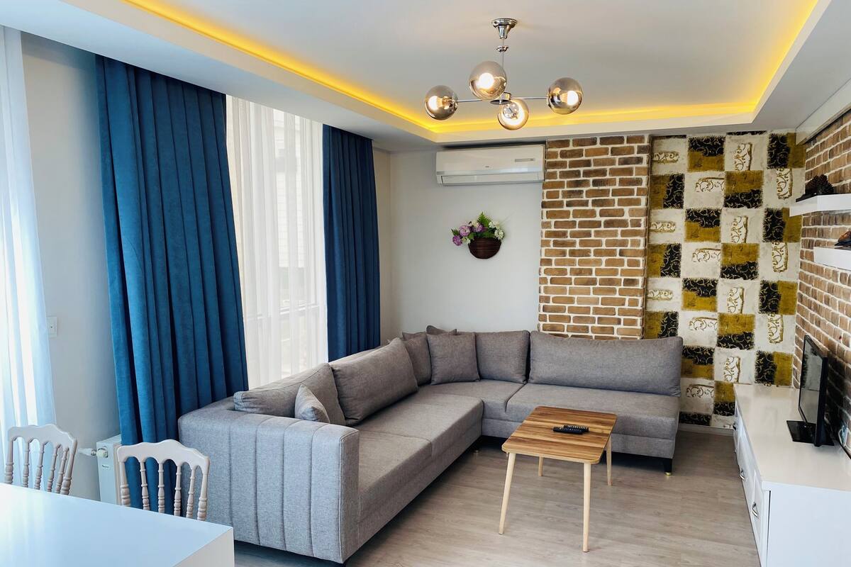 A modern living room with a grey sectional sofa, a wooden coffee table, a ceiling light fixture, and a brick-patterned wall.