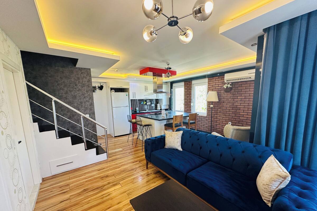 A modern living room with a blue sofa, wooden flooring, a staircase, and a kitchen in the background.
