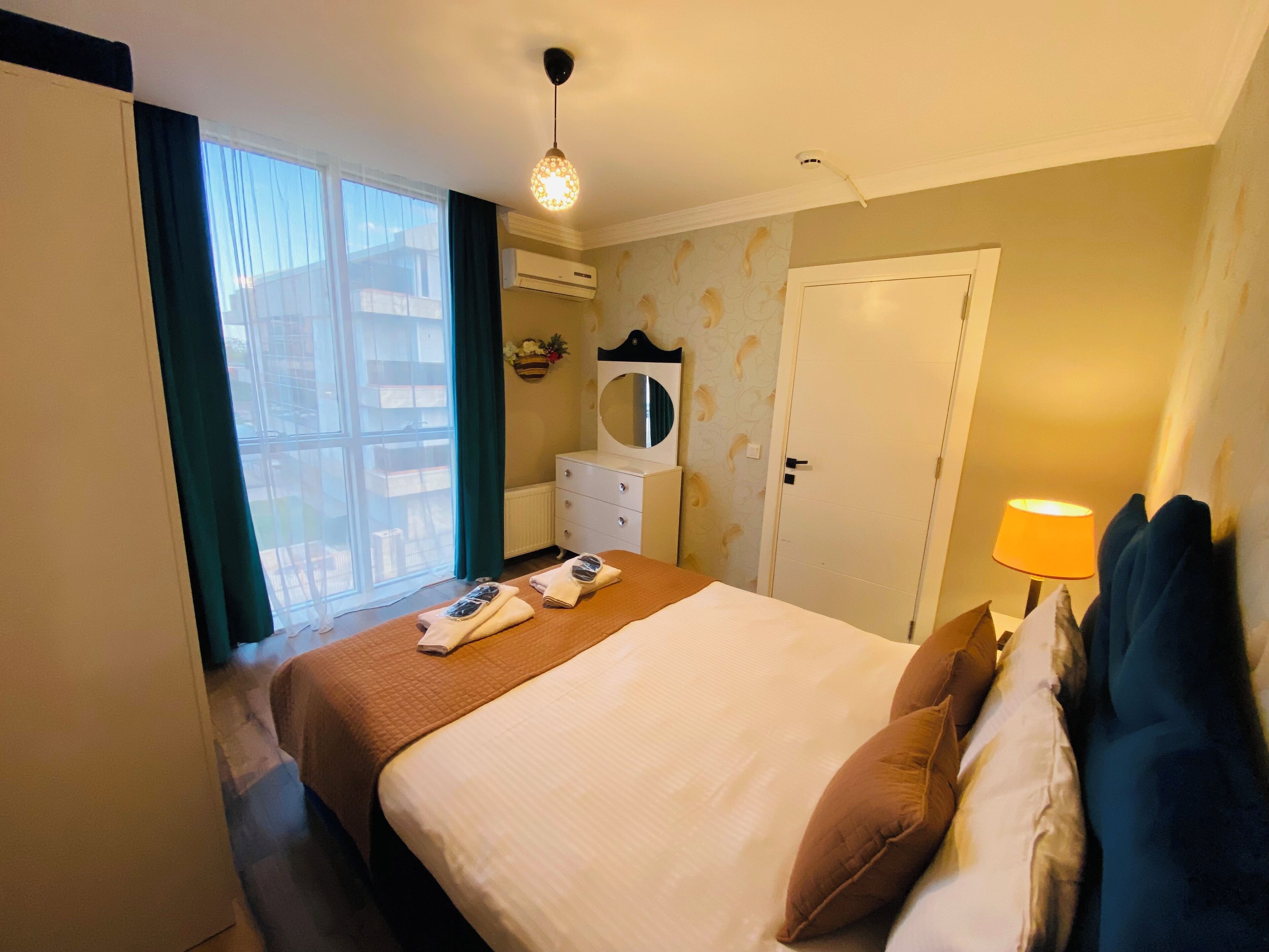 A hotel room with a bed, a bedside table with a lamp, a dresser, a mirror, and a window with curtains.