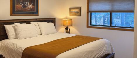 A bedroom with a wooden bed, white bedding, a lamp, two framed pictures, and a window with a view of trees.