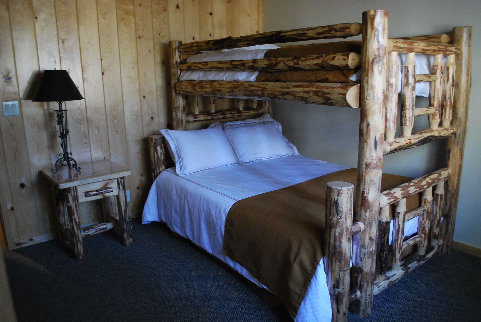 Uma cama beliche com estrutura de madeira, uma mesa de cabeceira com abajur e um chão coberto por carpete azul.
