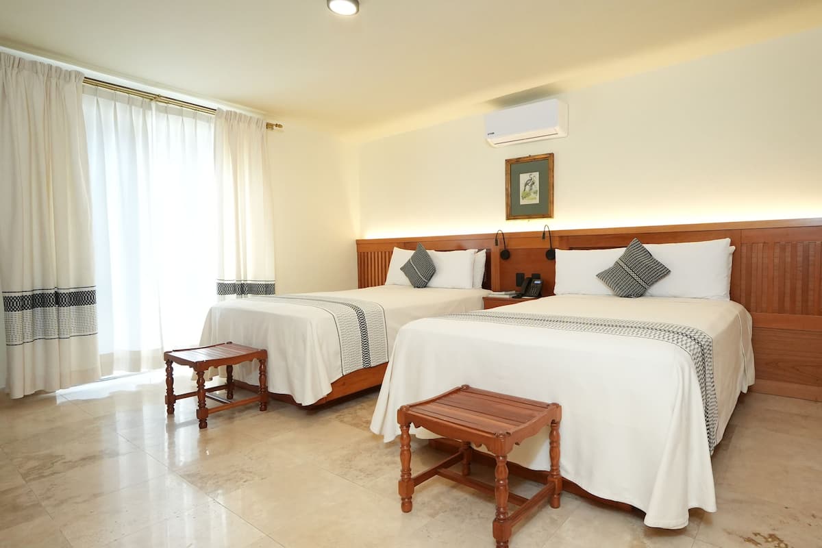Habitación de hotel con dos camas, cabecero de madera, mesita de noche, taburete pequeño de madera y un cuadro en la pared.
