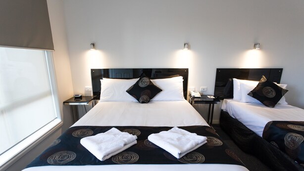 Ein Hotelzimmer mit zwei Betten, jeweils mit einer schwarz-weiß gemusterten Tagesdecke, weißen Kissen und einem schwarzen Kopfteil. Es gibt zwei Nachttische mit Lampen, ein Fenster mit Jalousie und ein großes Fenster auf der linken Seite.