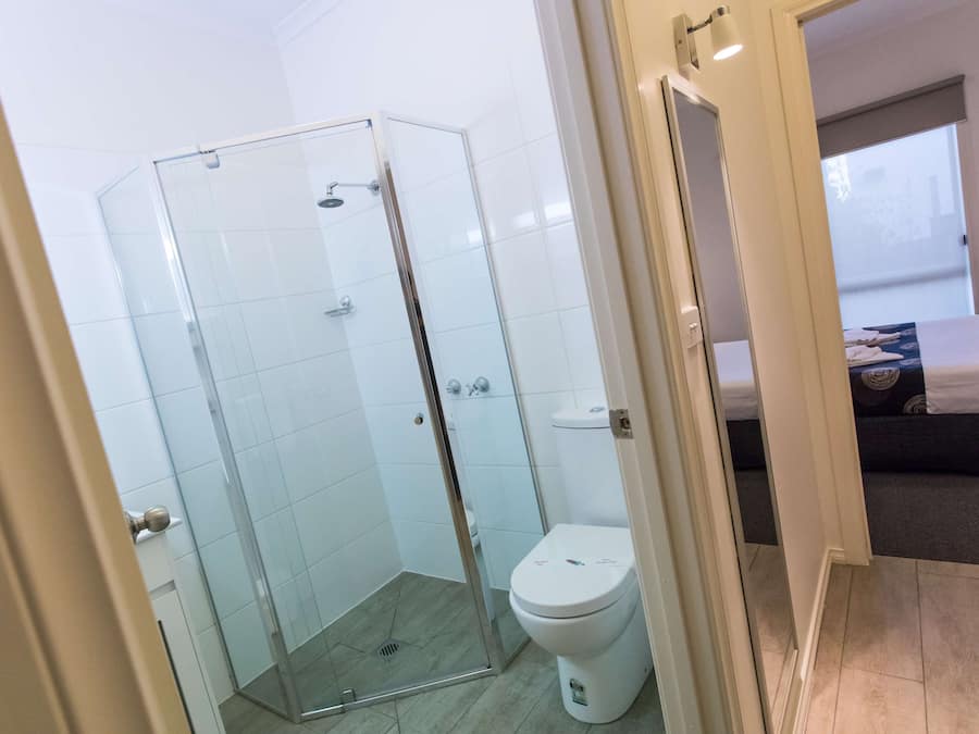 Un bagno moderno con cabina doccia in vetro, un water bianco e un letto visibile nella stanza adiacente.