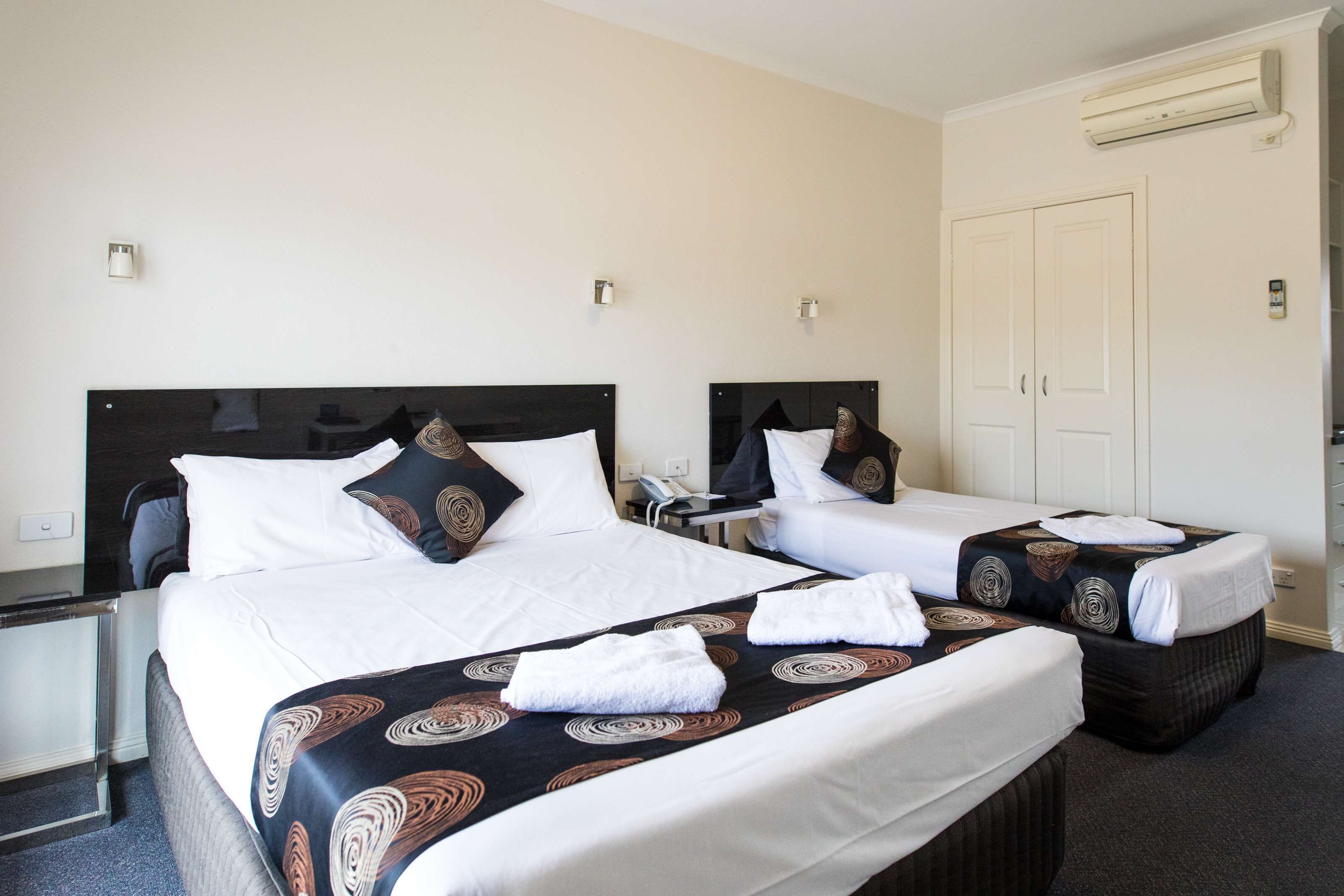 Quarto de hotel com duas camas, cada uma com cabeceira, roupa de cama branca e colcha estampada. Há uma mesa de cabeceira com uma luminária, telefone e uma mesinha. O quarto possui uma porta e um aparelho de ar condicionado na parede.