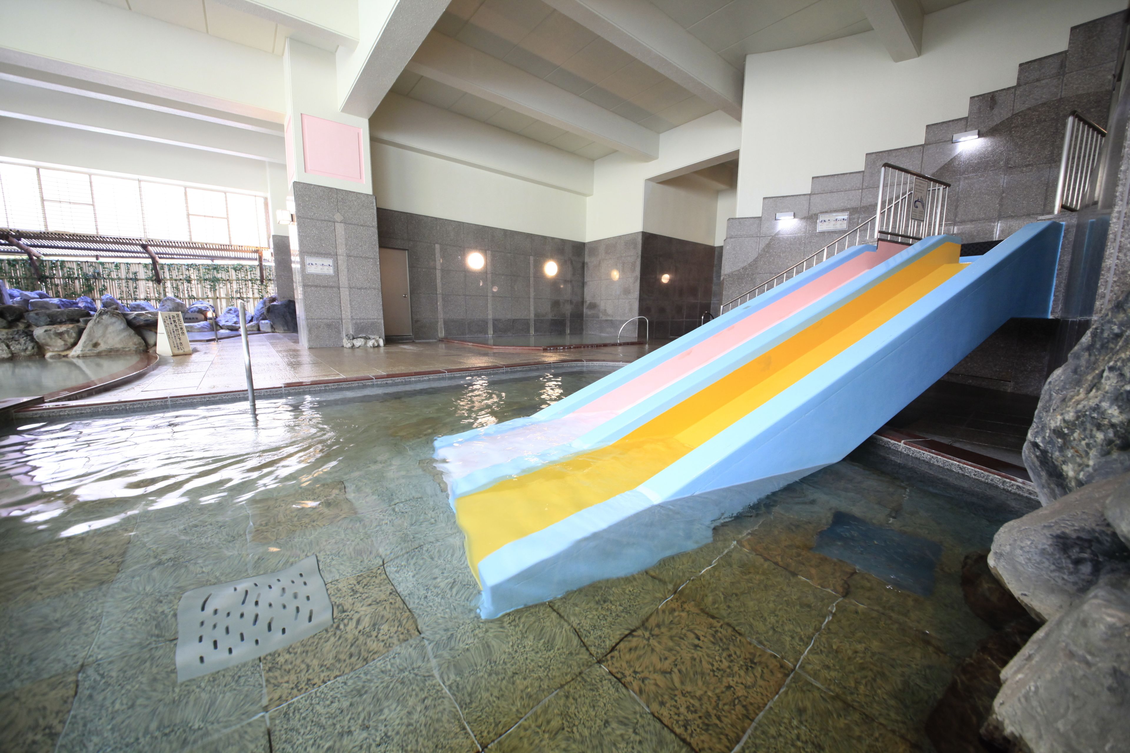 An indoor water slide with a blue, yellow, and pink section, surrounded by stone walls and a tiled floor.