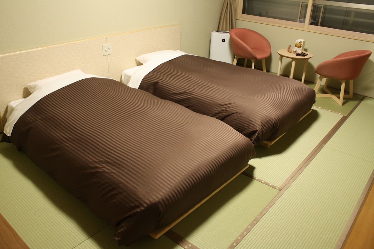 A hotel room with two single beds, a small table with a chair, and a window with curtains.