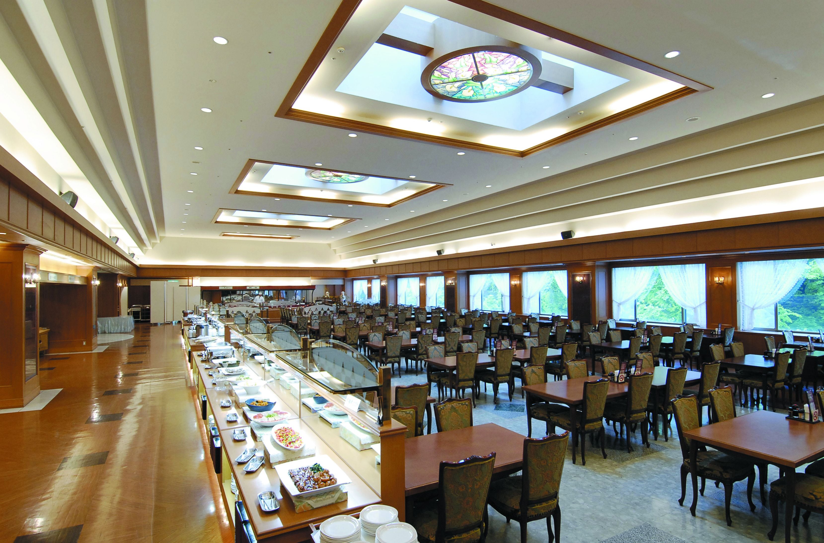 A spacious hotel dining area with multiple tables and chairs, a buffet line, and large windows offering views of the outdoors.