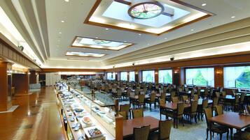 Amplia zona de comedor de hotel con numerosas mesas y sillas, una barra de buffet y grandes ventanales que ofrecen vistas al exterior.