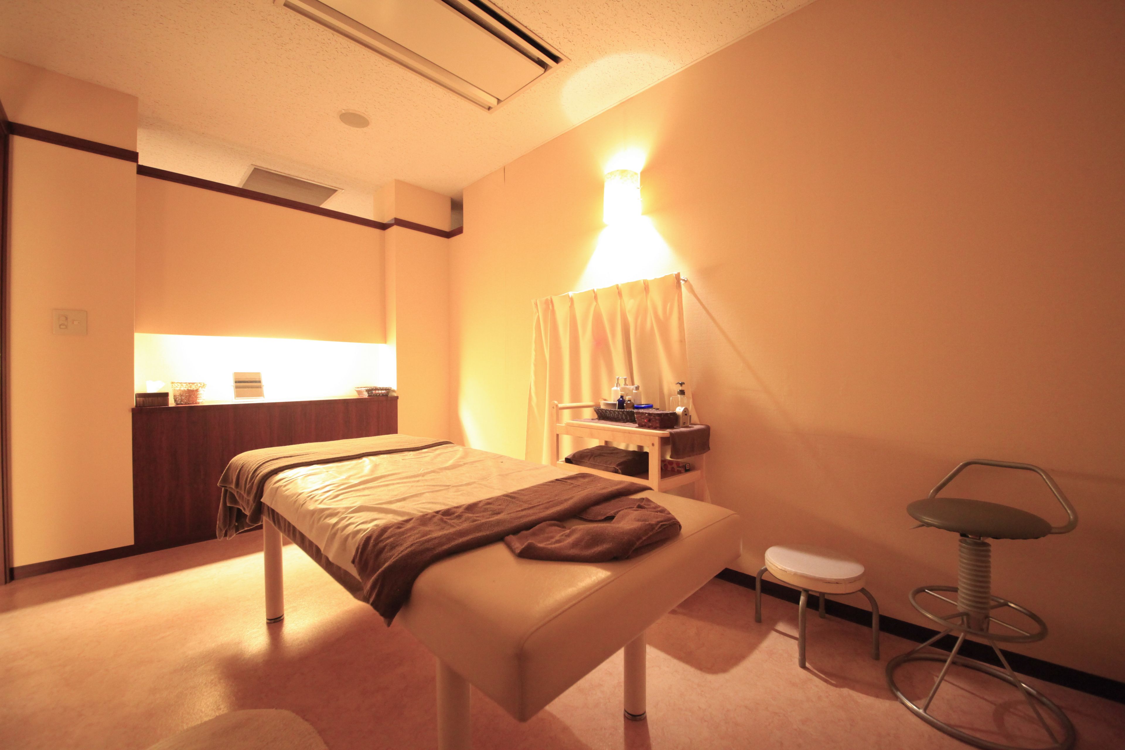 A hotel room with a bed, a bedside table, a small stool, a lamp, and a wall-mounted shelf.
