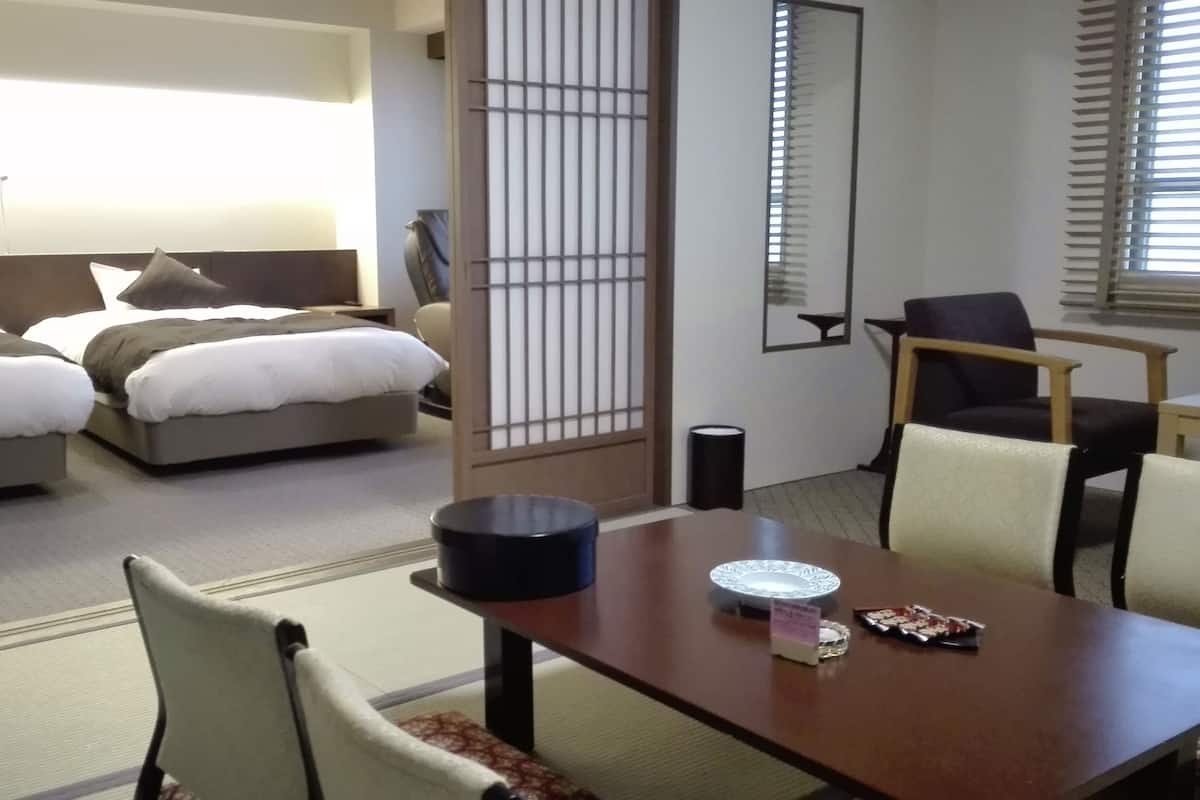 A hotel room with a bed, a desk, and a chair. There is a window with blinds, a mirror, and a small trash can.