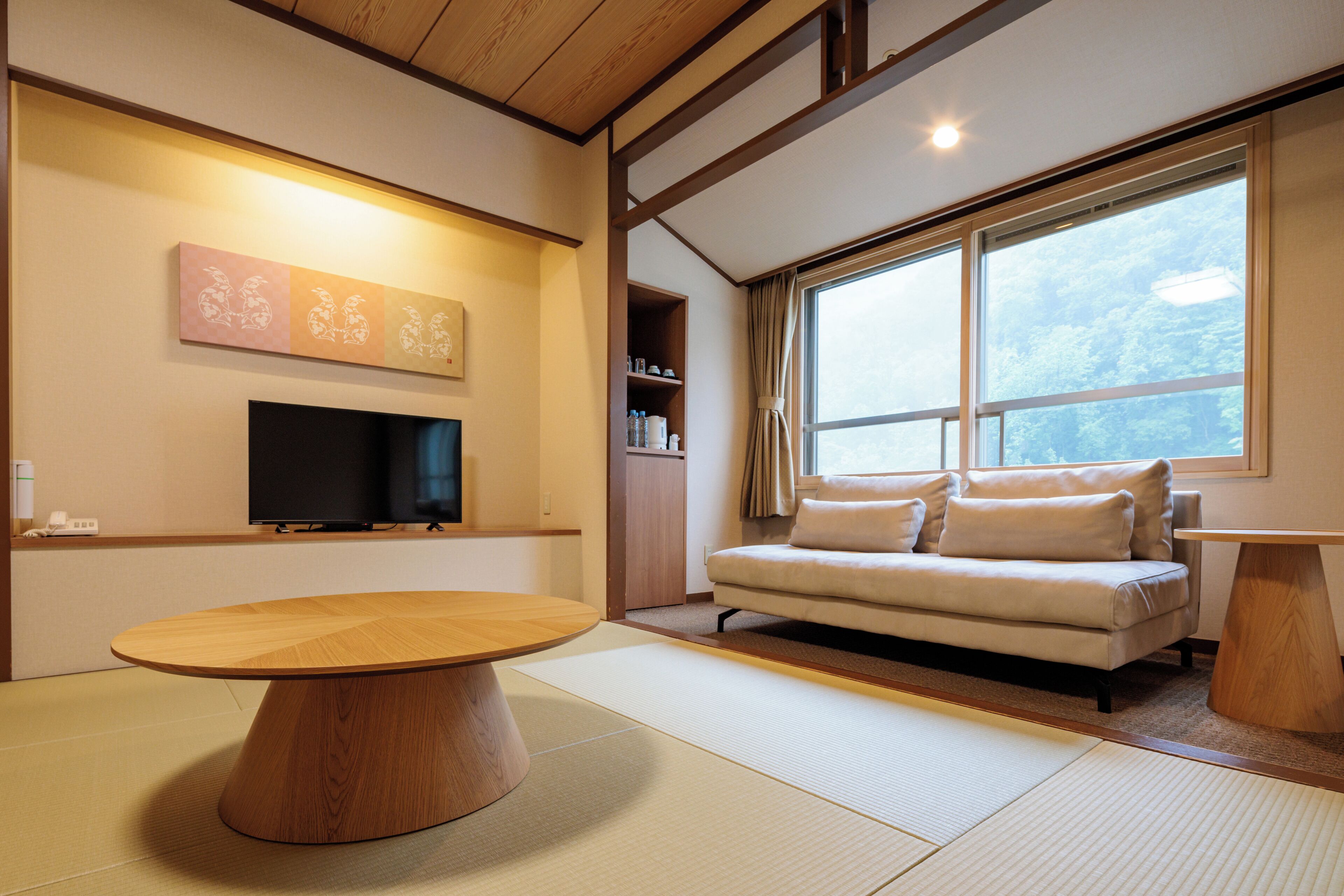 A modern living room with a wooden dining table, a sofa, and a large window providing a view of the outdoors.