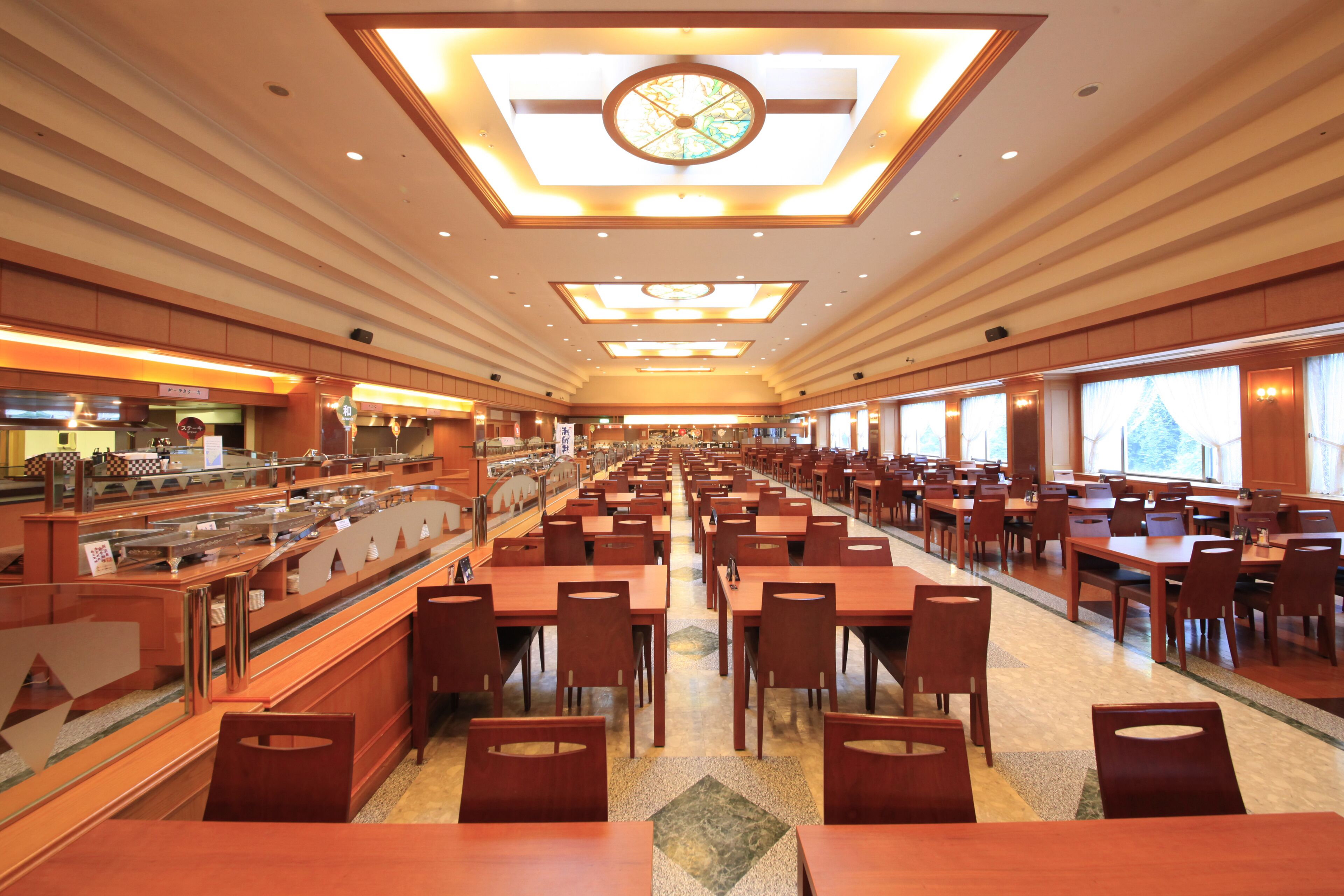 A spacious hotel dining area with multiple tables and chairs, a buffet counter, and large windows providing natural light.