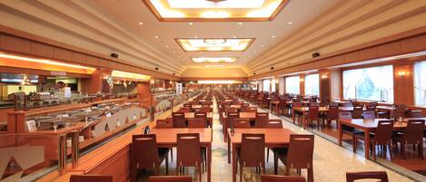 A spacious hotel dining area with multiple tables and chairs, a buffet counter, and large windows providing natural light.
