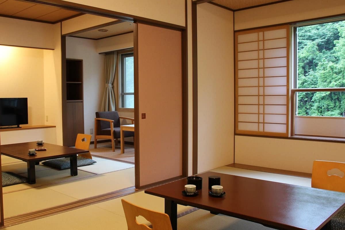 Une pièce de style japonais traditionnel, avec un sol recouvert de tatamis, une table basse avec des coussins, un téléviseur et une fenêtre donnant sur un paysage verdoyant.