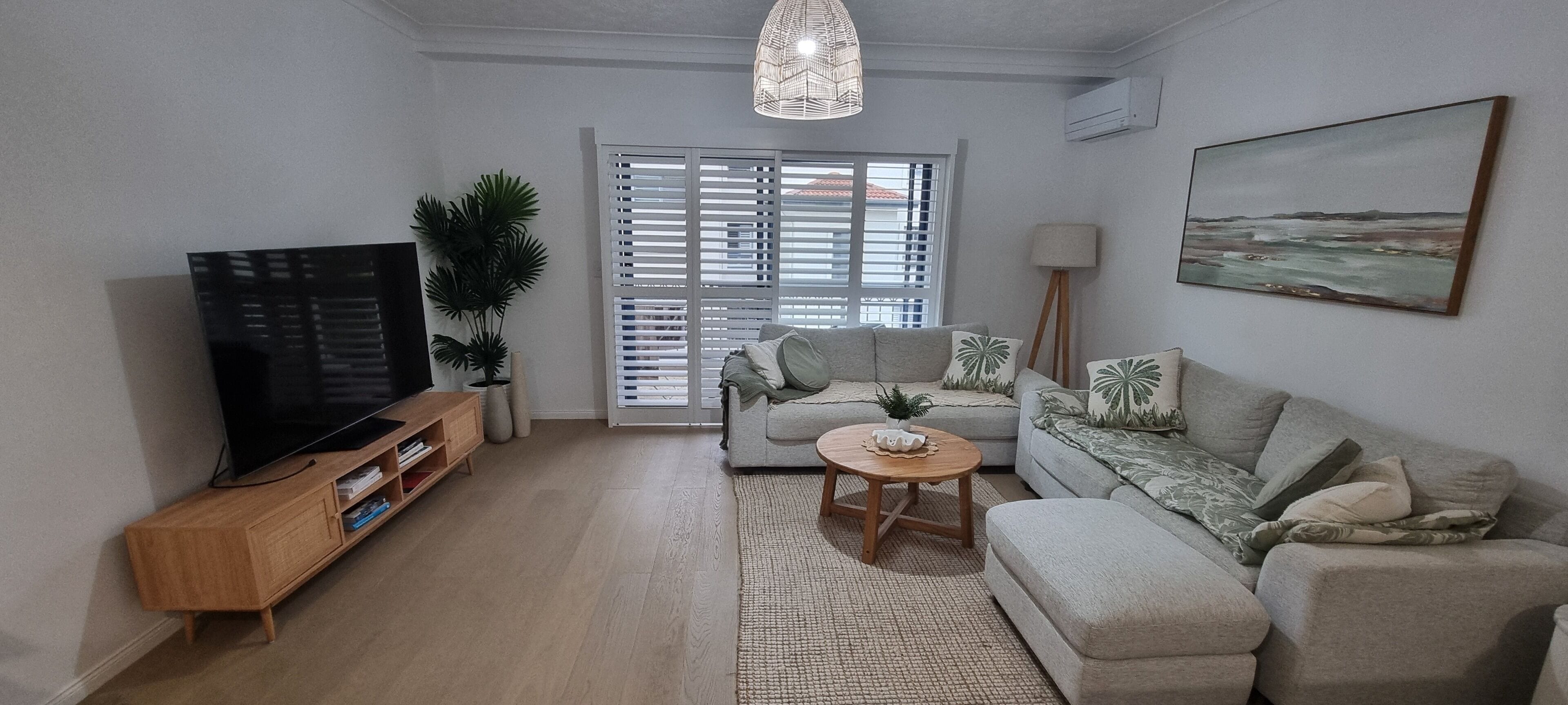 A living room with a sofa, a coffee table, a TV on a wooden stand, and a large window with blinds.