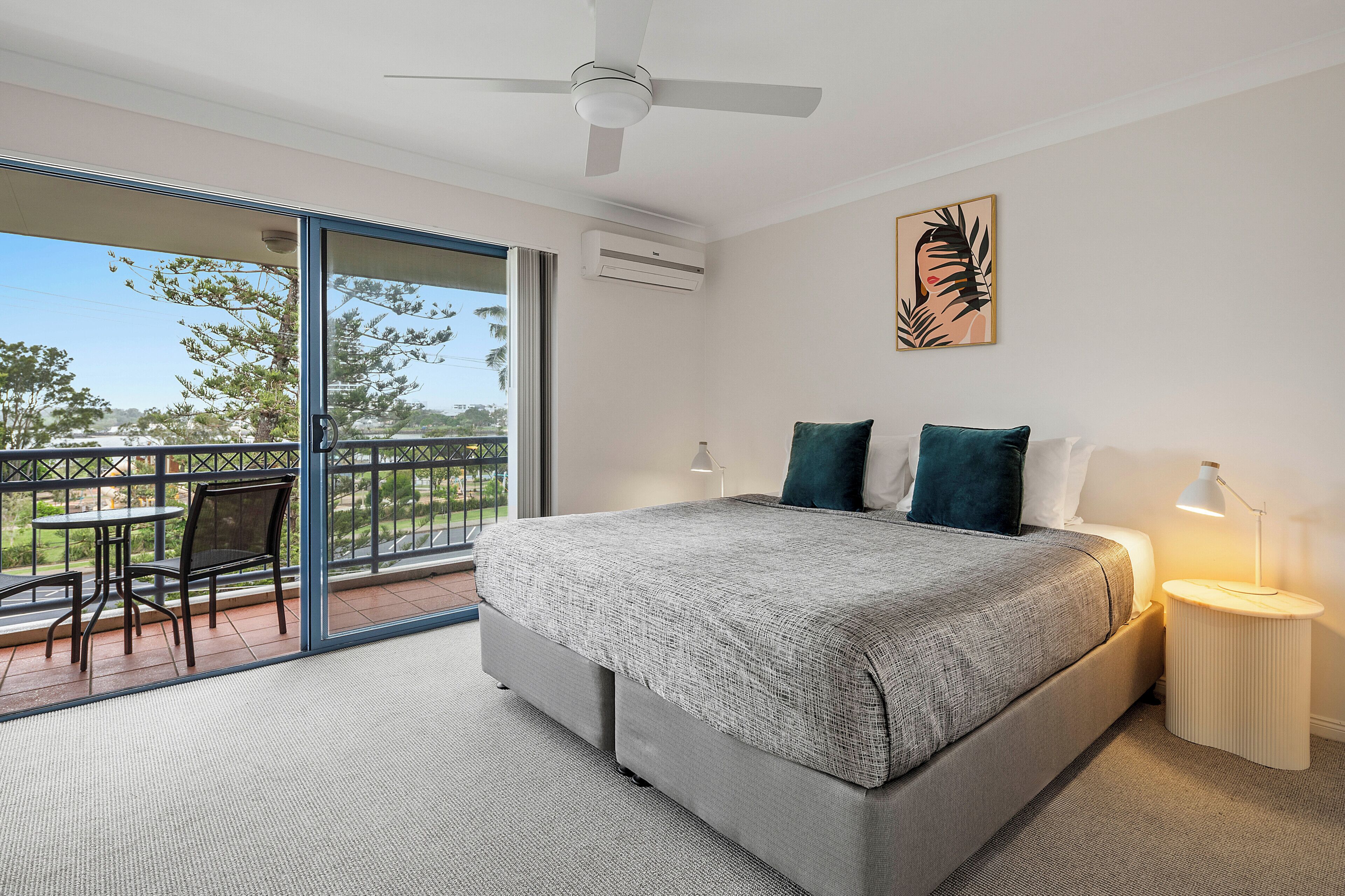 Un dormitorio con una cama grande, un ventilador de techo, una mesita de noche con lámpara, una ventana con vista al exterior y un cuadro en la pared.
