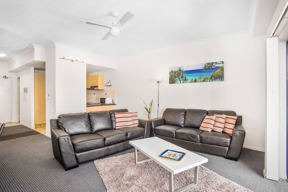 A modern living room with two leather sofas, a white coffee table, and a painting of a tropical beach.