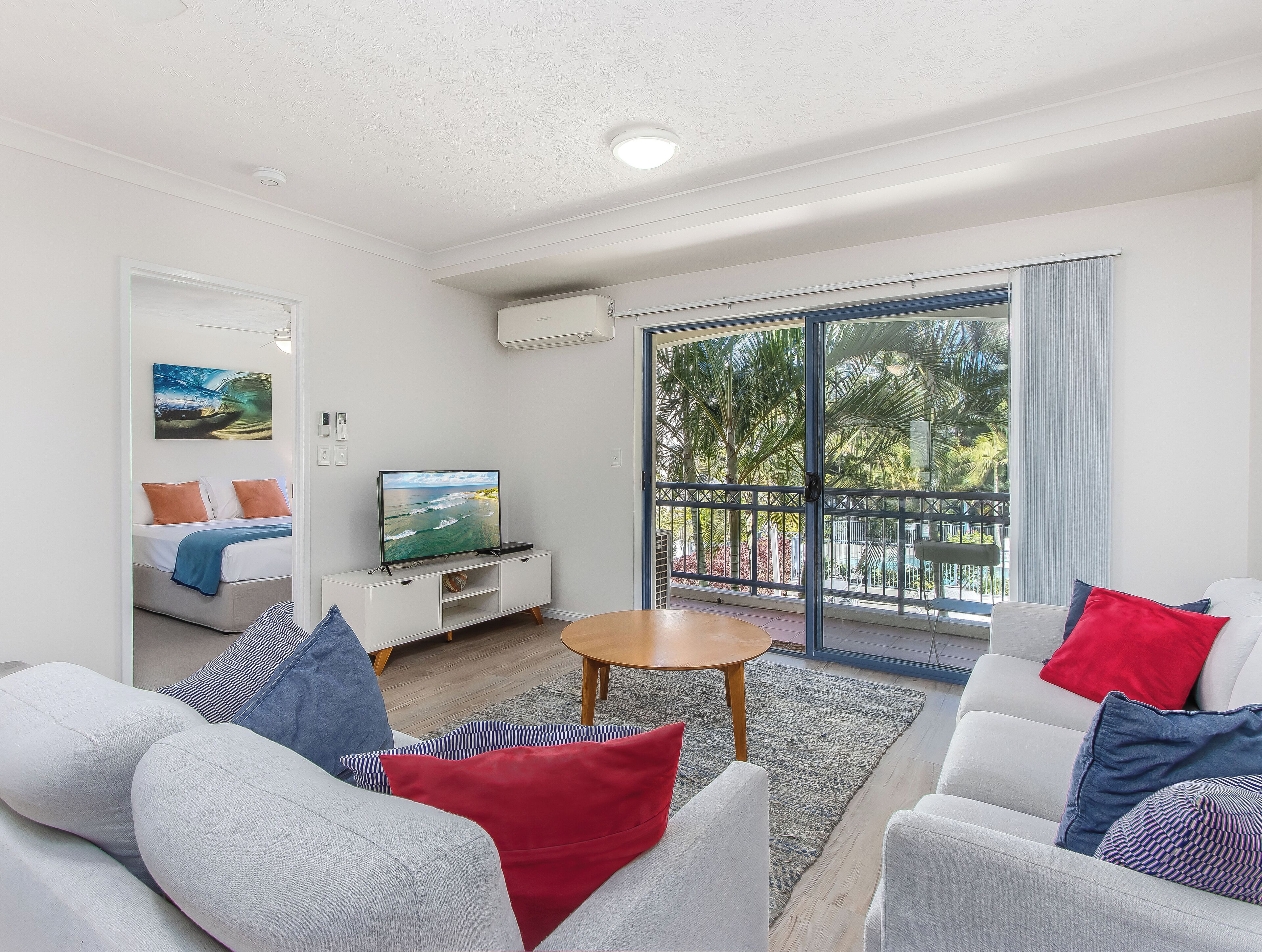 A modern living room with a sofa set, a TV on a stand, and a balcony with a view of palm trees.