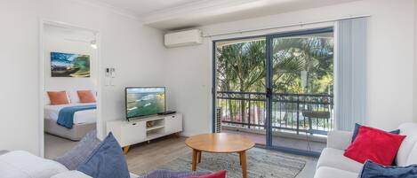 A modern living room with a sofa set, a TV on a stand, and a balcony with a view of palm trees.