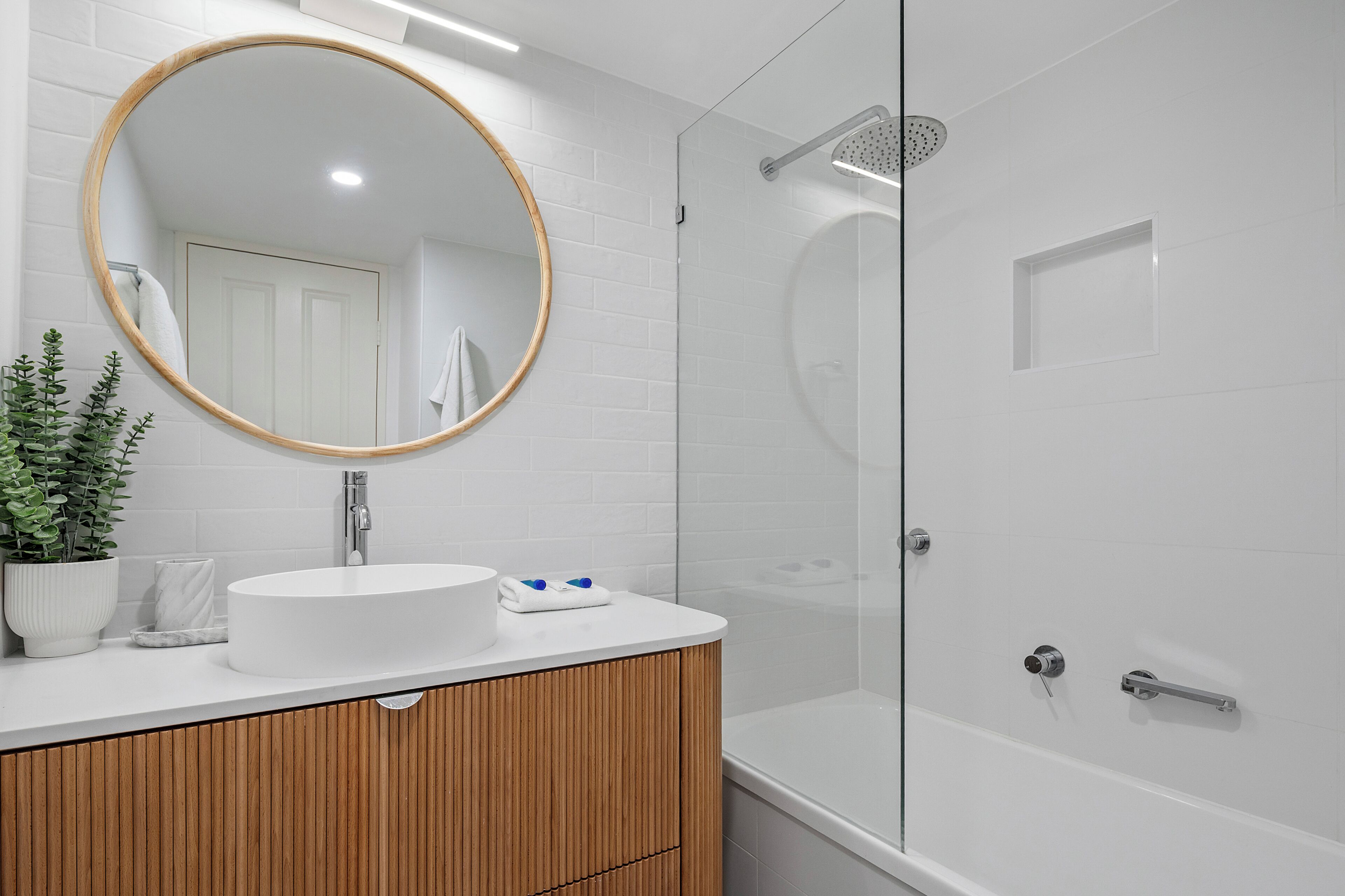 A modern bathroom with a round mirror, white sink, wooden vanity, glass shower enclosure, and a white door.