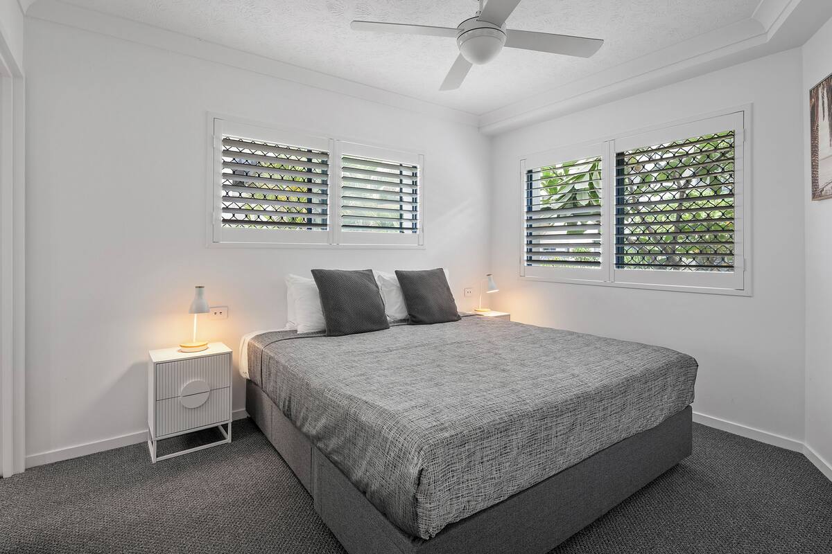 A bedroom with a bed, a nightstand, a lamp, a ceiling fan, and windows with blinds.