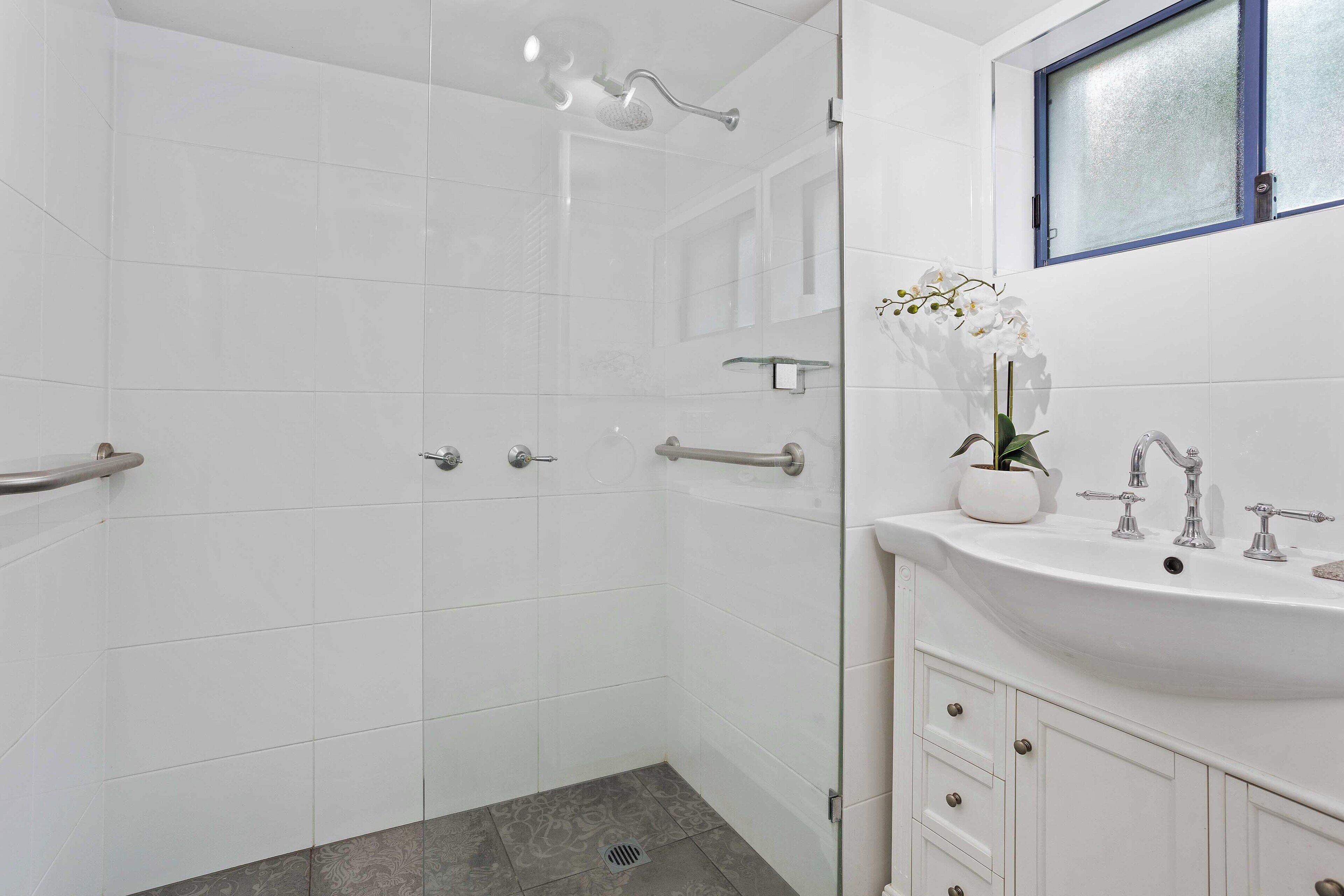 A modern bathroom with a glass shower enclosure, a white sink with a vase of flowers, and a window above the sink.