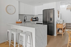 A modern kitchen with white cabinetry, a stainless steel refrigerator, a marble island with bar stools, and a microwave. - Bella Mare Beachside Apartments (Coolangatta)