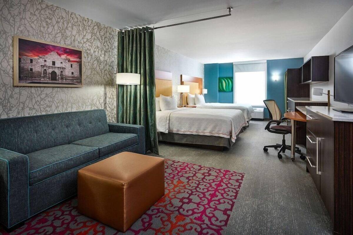 Ein Hotelzimmer mit Bett, Sofa, Schreibtisch, Fernseher und einem Gemälde eines Gebäudes.