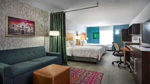 A hotel room with a bed, a sofa, a desk, a TV, and a painting on the wall. - Home2 Suites by Hilton San Antonio Airport, TX (San Antonio)