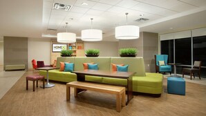 A modern hotel lobby with a green sofa, wooden table, and chairs. There are potted plants and a sign that reads "HOME2". - Home2 Suites by Hilton San Antonio Airport, TX (San Antonio)