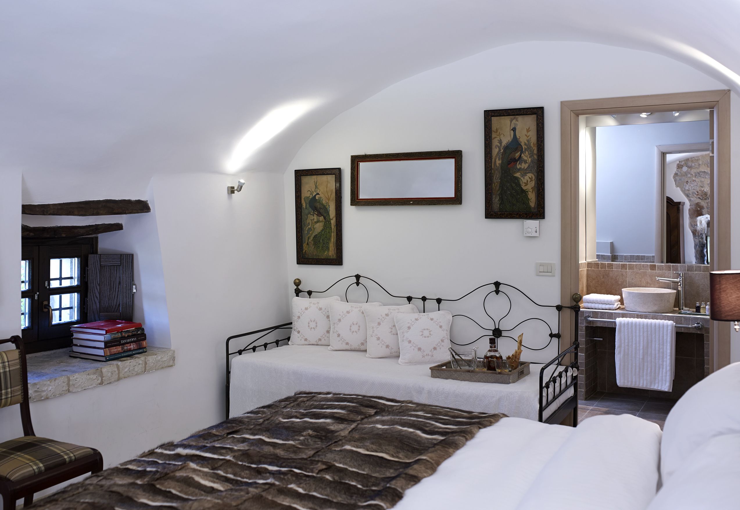 Un dormitorio con una cama, una mesita de noche con libros, un espejo y un baño visible a través de una puerta abierta.