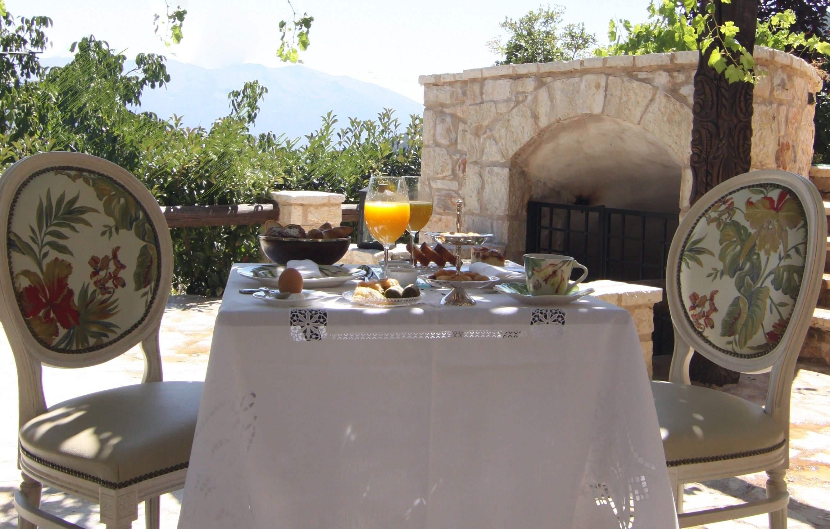 A table set outdoors with a stone fireplace, surrounded by chairs with floral upholstery.